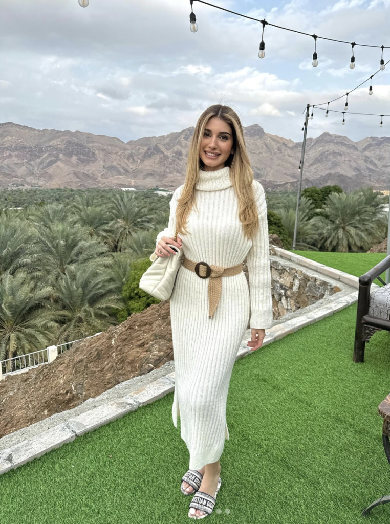 A woman in a white sweater dress, posing outdoors.